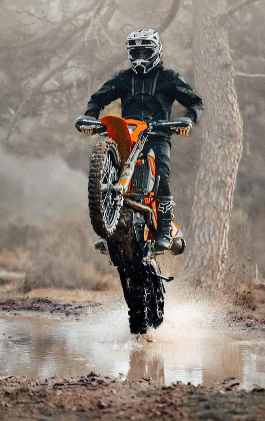 Person riding a dirt bike through a muddy puddle with a forest background
